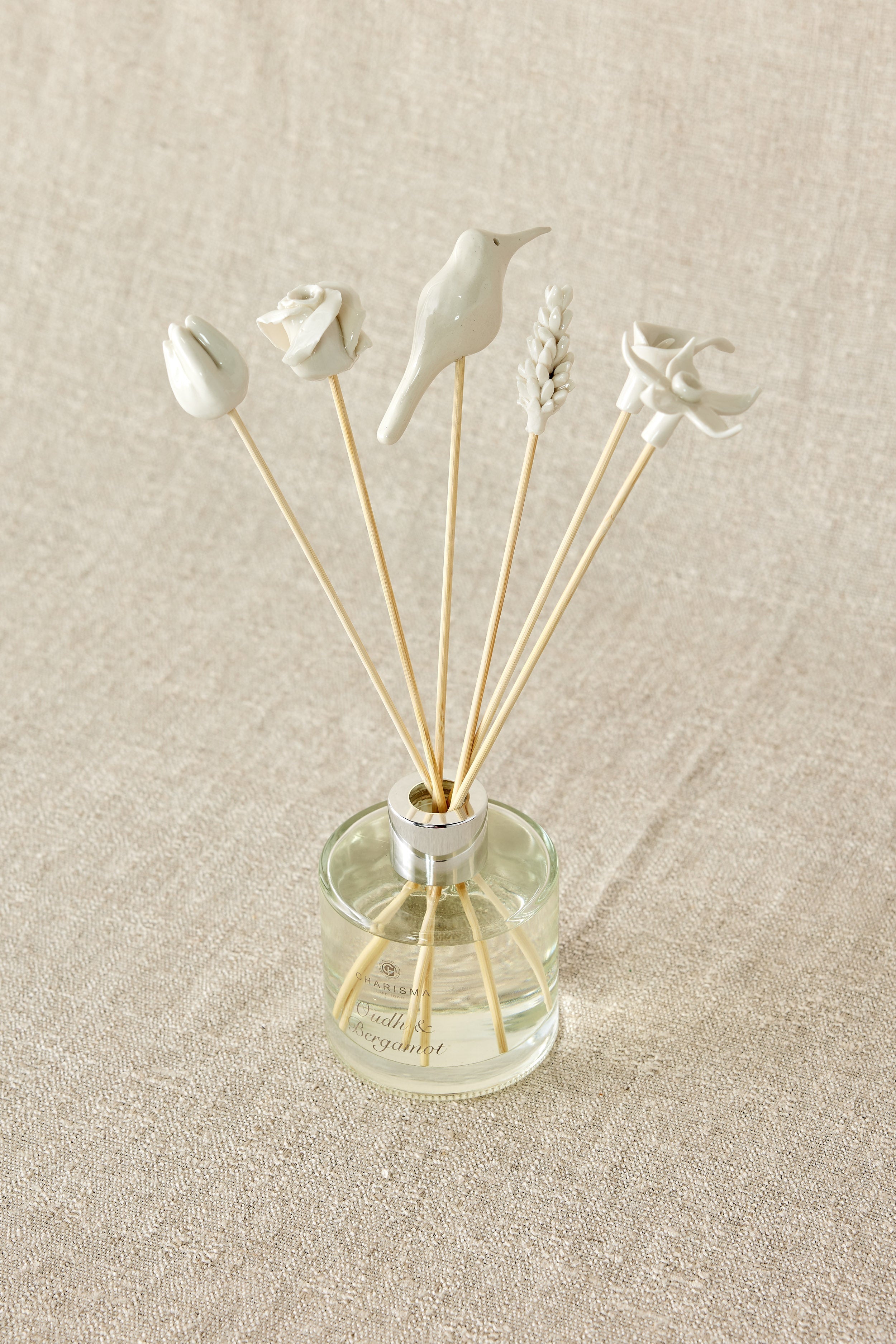 Decorative diffuser with bird-shaped sticks in a clear bottle on a beige fabric background
