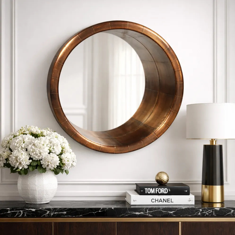 Round copper framed mirror on a wall above a console table with decor items.