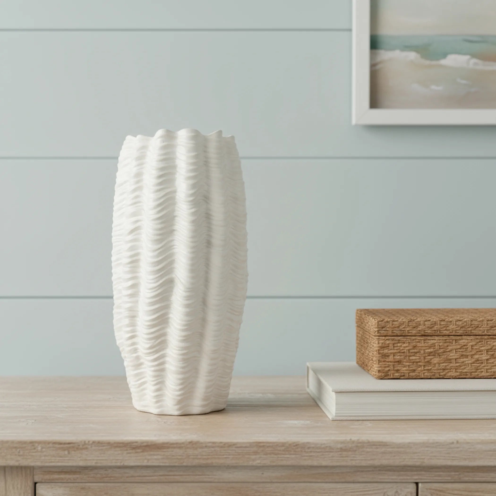 White textured vase on a wooden surface with a light blue wall and framed picture in the background