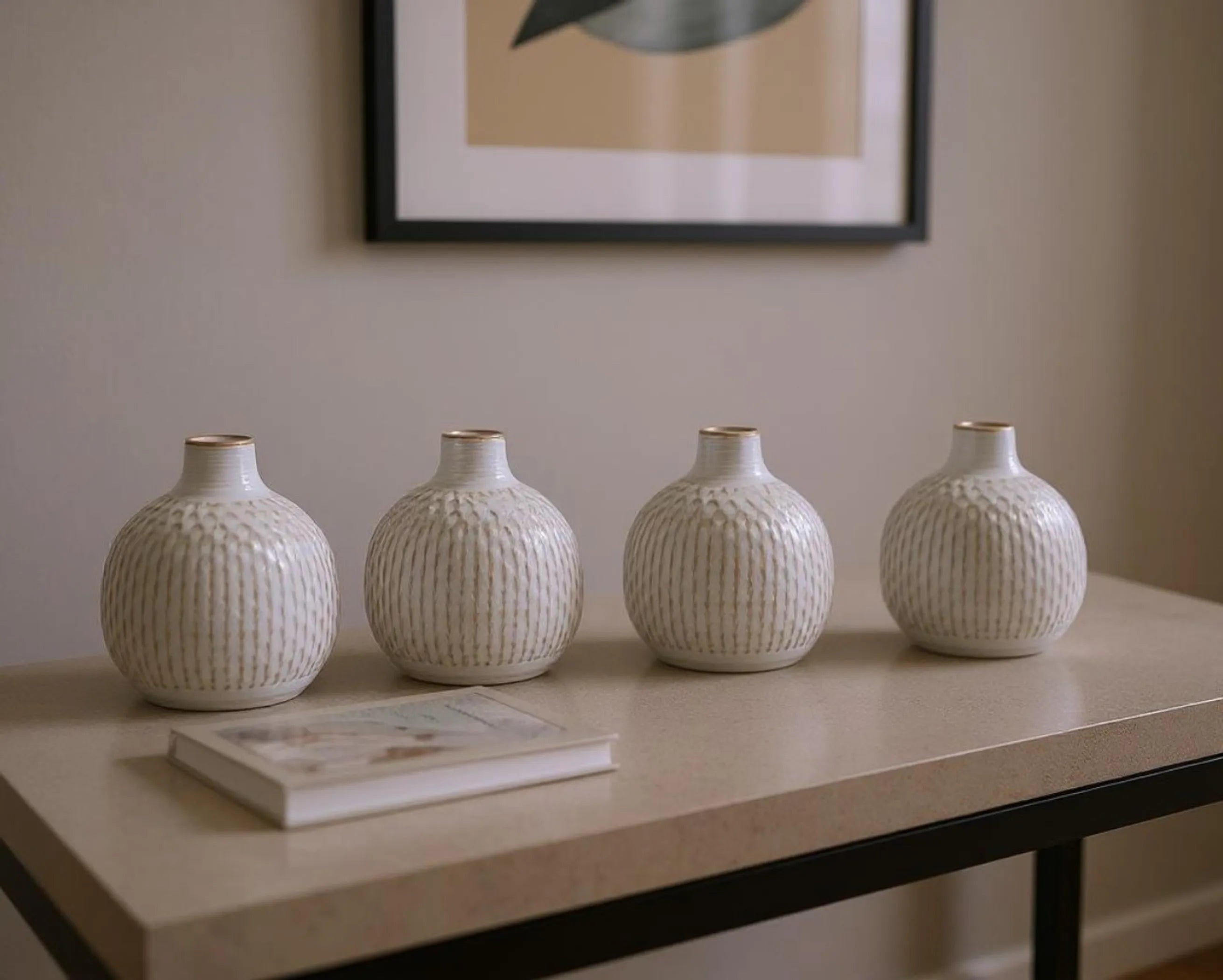 Four white textured vases on a table with a framed picture in the background.