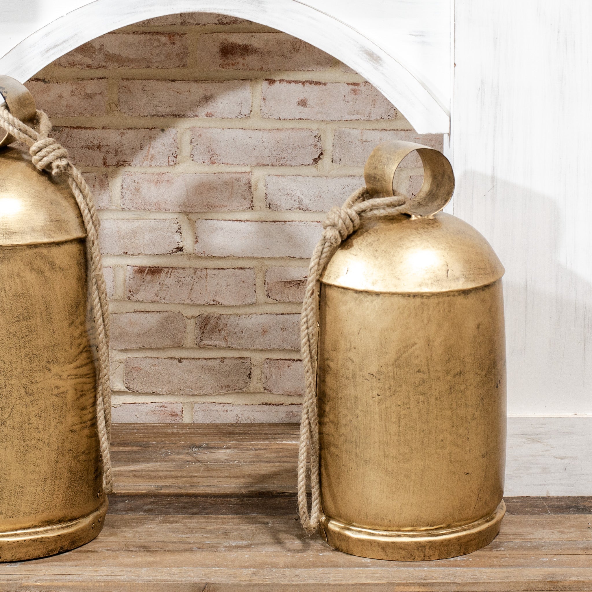 Two gold decorative holiday bells items with ropes against a brick wall.
