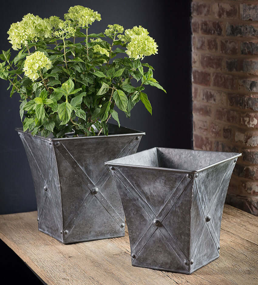 Two metallic planters with a green plant against a brick wall.