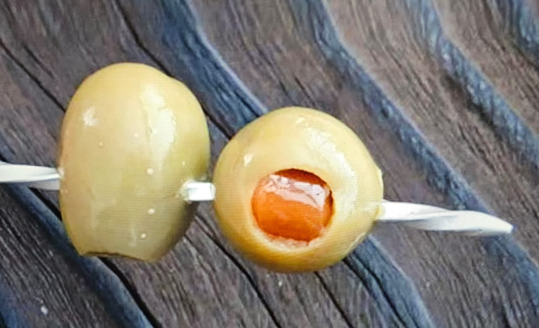 Two green olives with a silver plated swirl design martini cocktail stick between them on a wooden surface