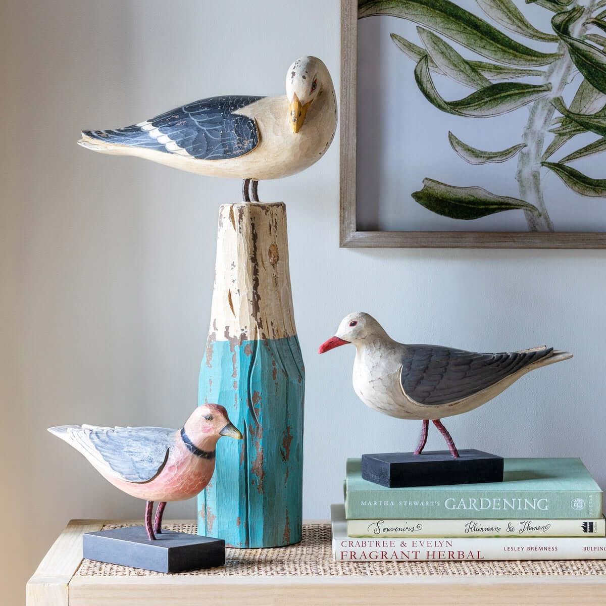 Decorative seagull figurines on books with a framed plant print in the background.