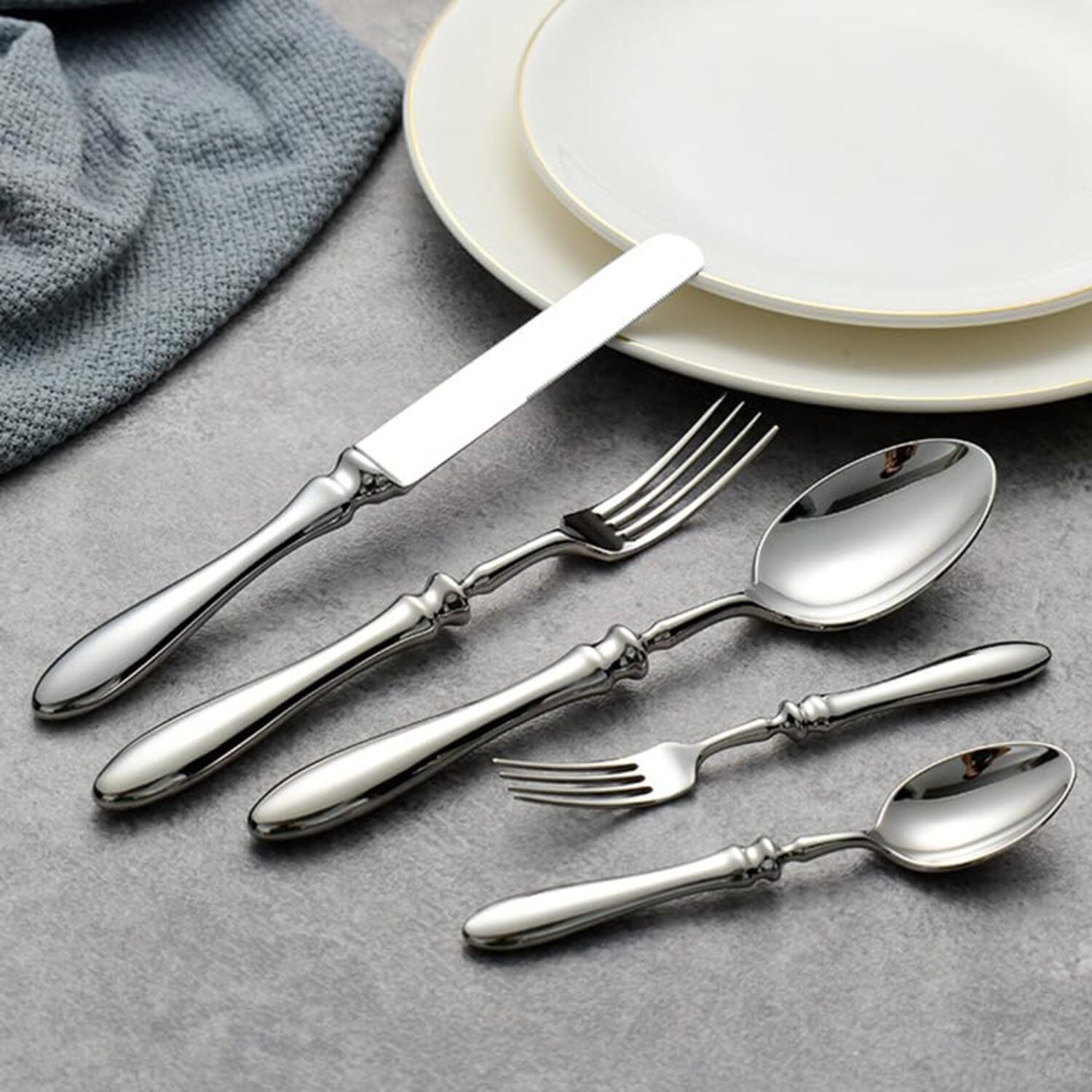 Set of silver cutlery including knives, forks, and spoons on a gray surface with plates in the background.