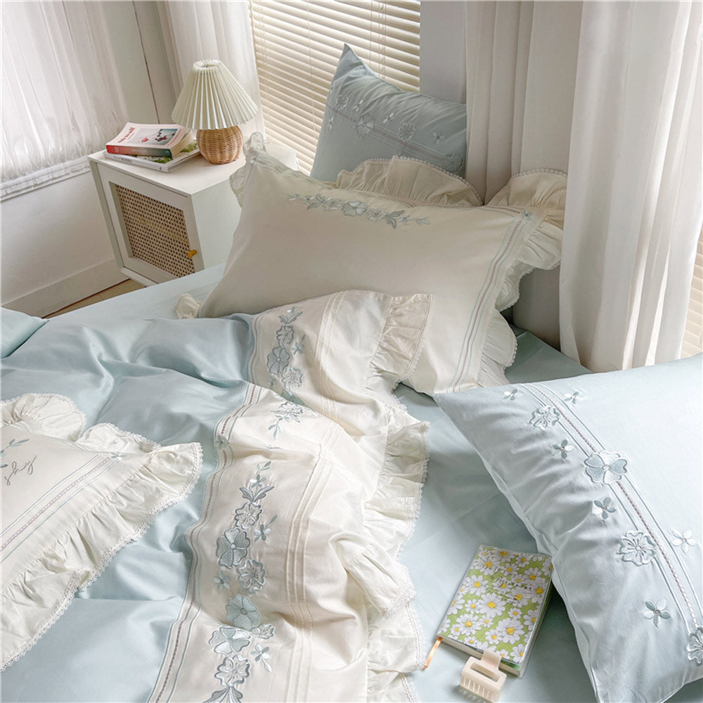Delicate embroidered bedding set on a bed with a side table and lamp in a softly lit room.