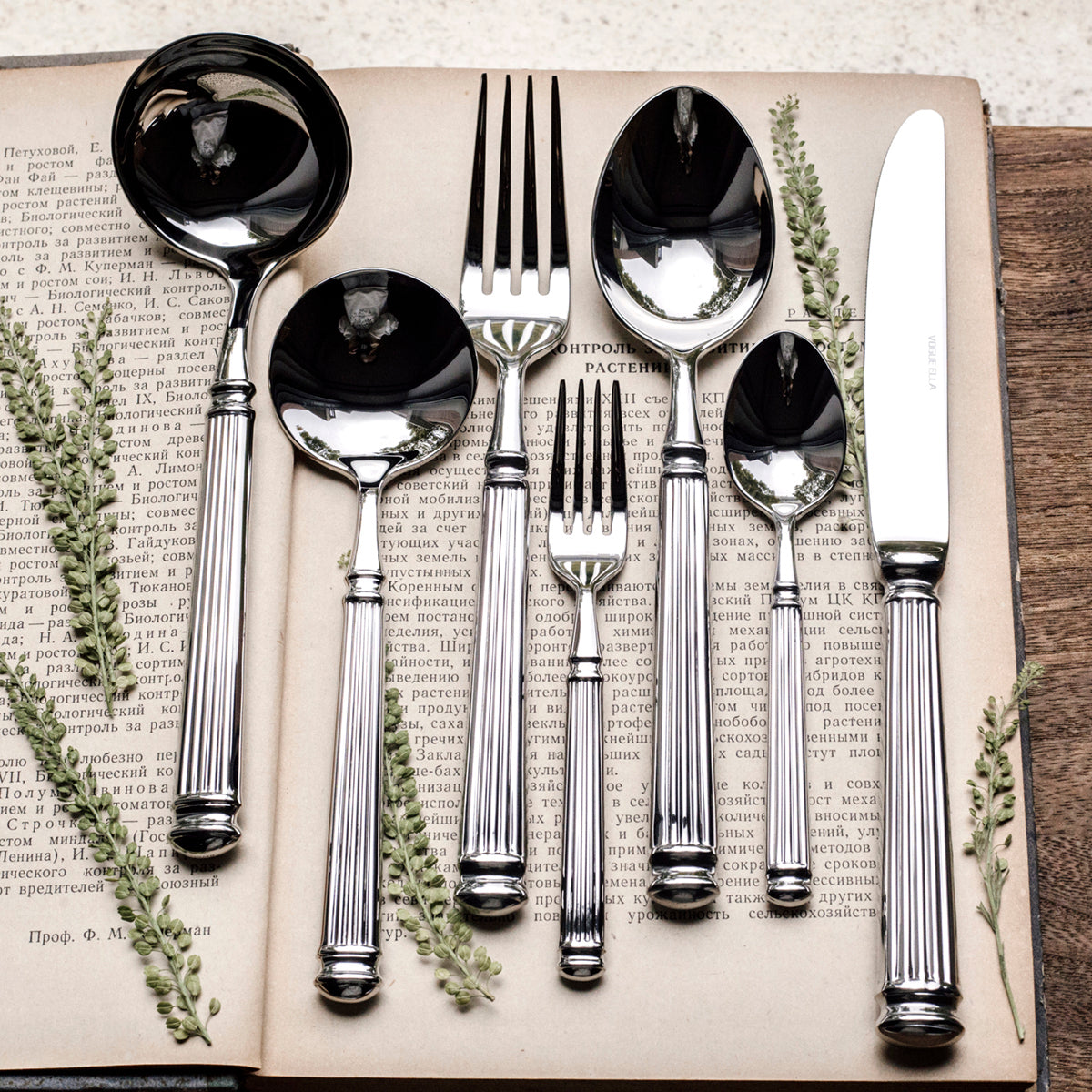 Set of silver cutlery including spoons, forks, and knives on an open book with greenery.