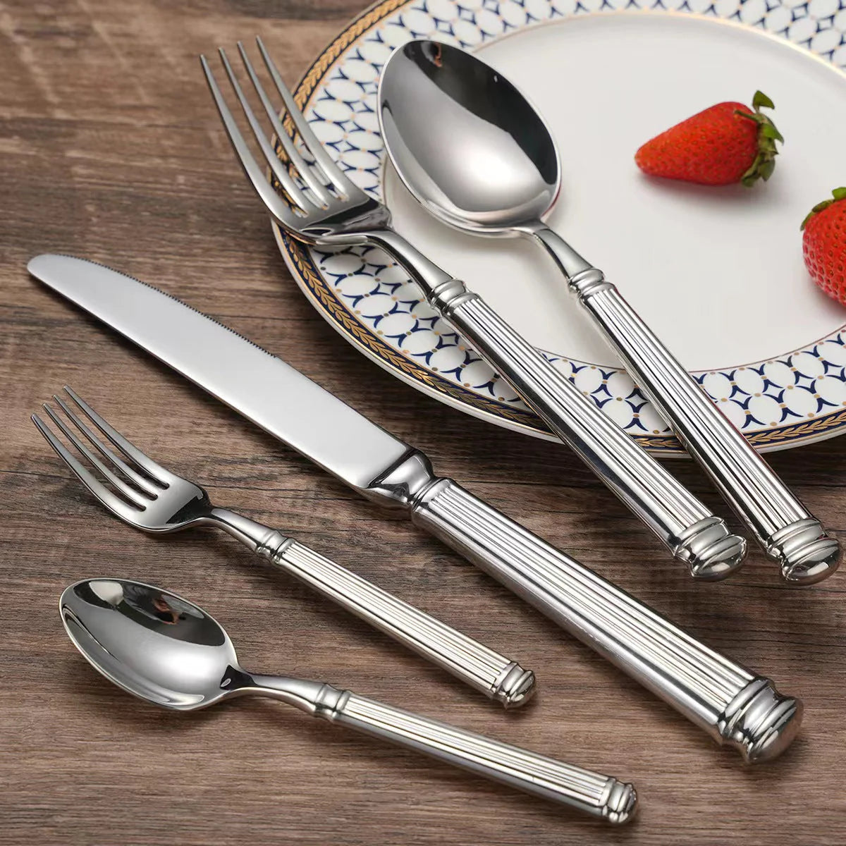 Silver cutlery set on a wooden table with a plate of strawberries in the background.