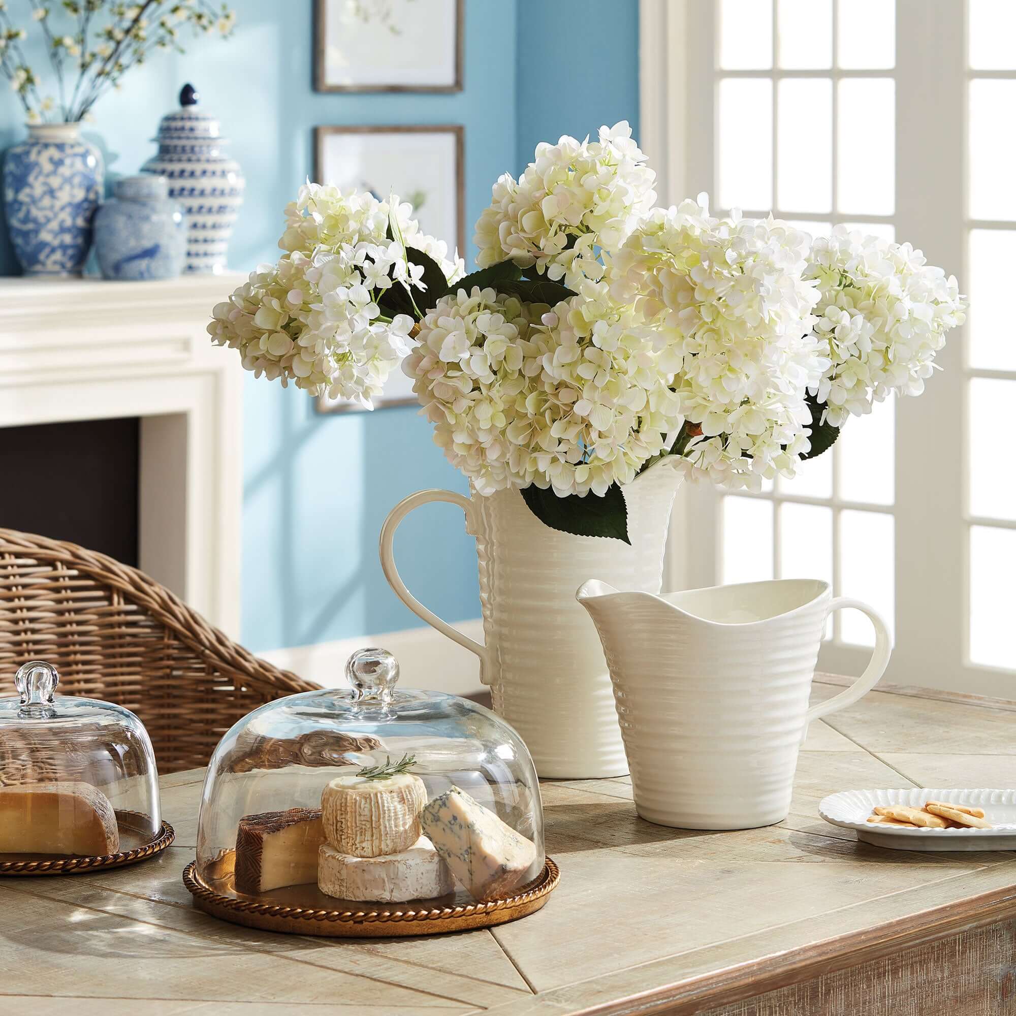 White flowers in a vase on a wooden table with a glass cloche and cheese platter.