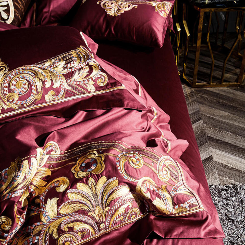 Luxurious burgundy bedding set with gold embroidery on a wooden floor.