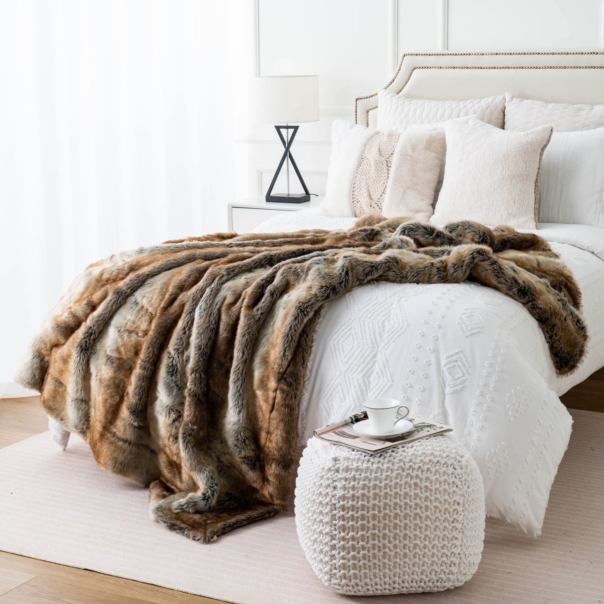 Bed with a fur throw blanket and a white ottoman with a cup on a light wood floor.