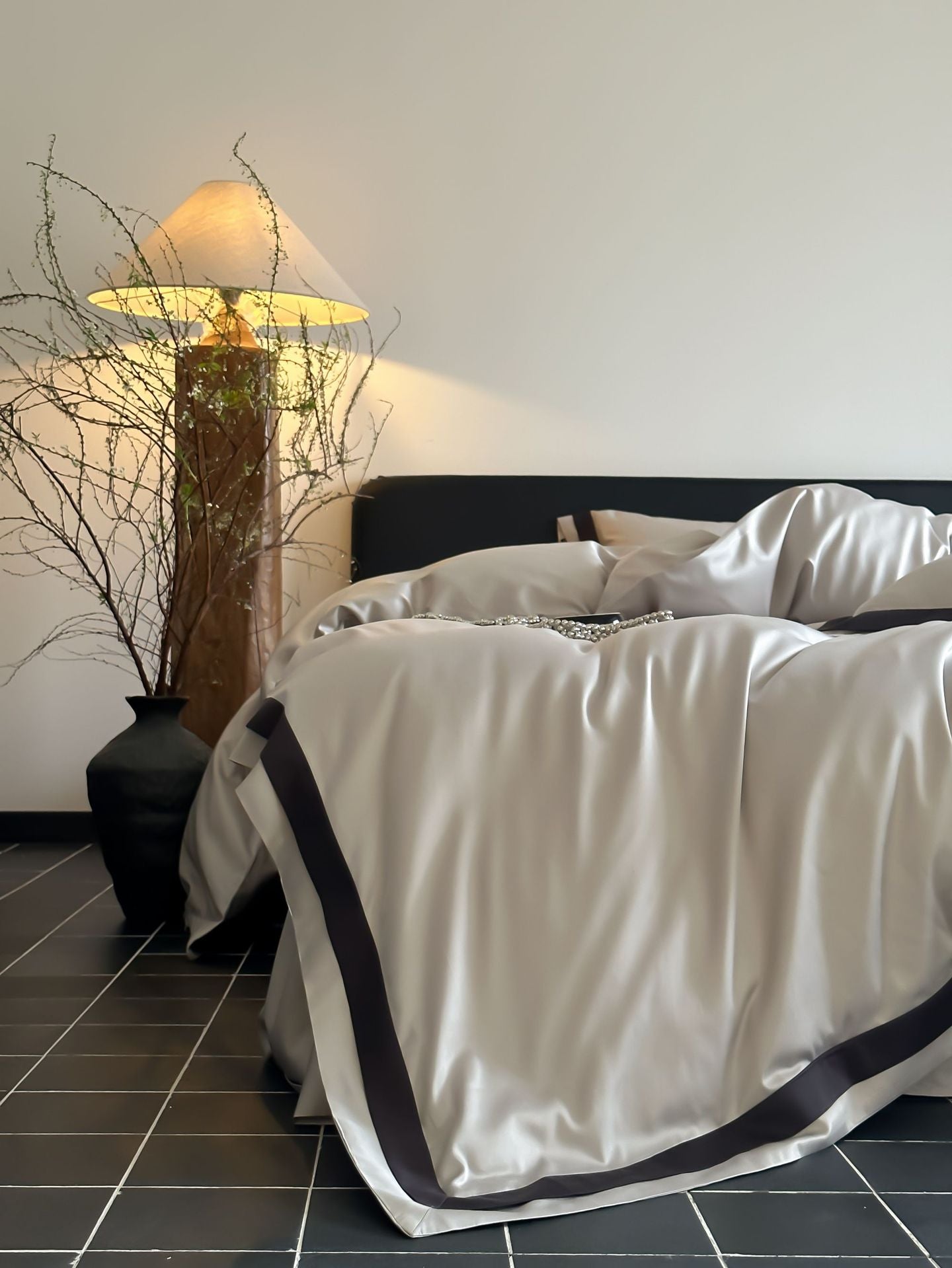 Bed with white bedding and black trim in a room with a lamp and decorative plant.