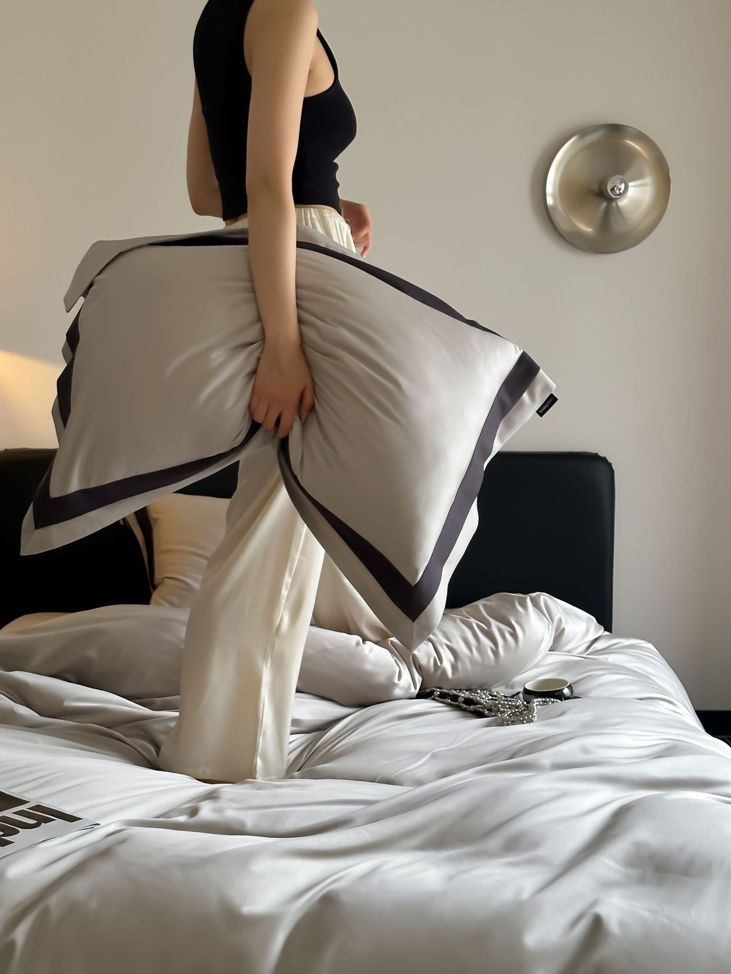 Person holding a white towel with black trim over a bed in a bedroom setting.