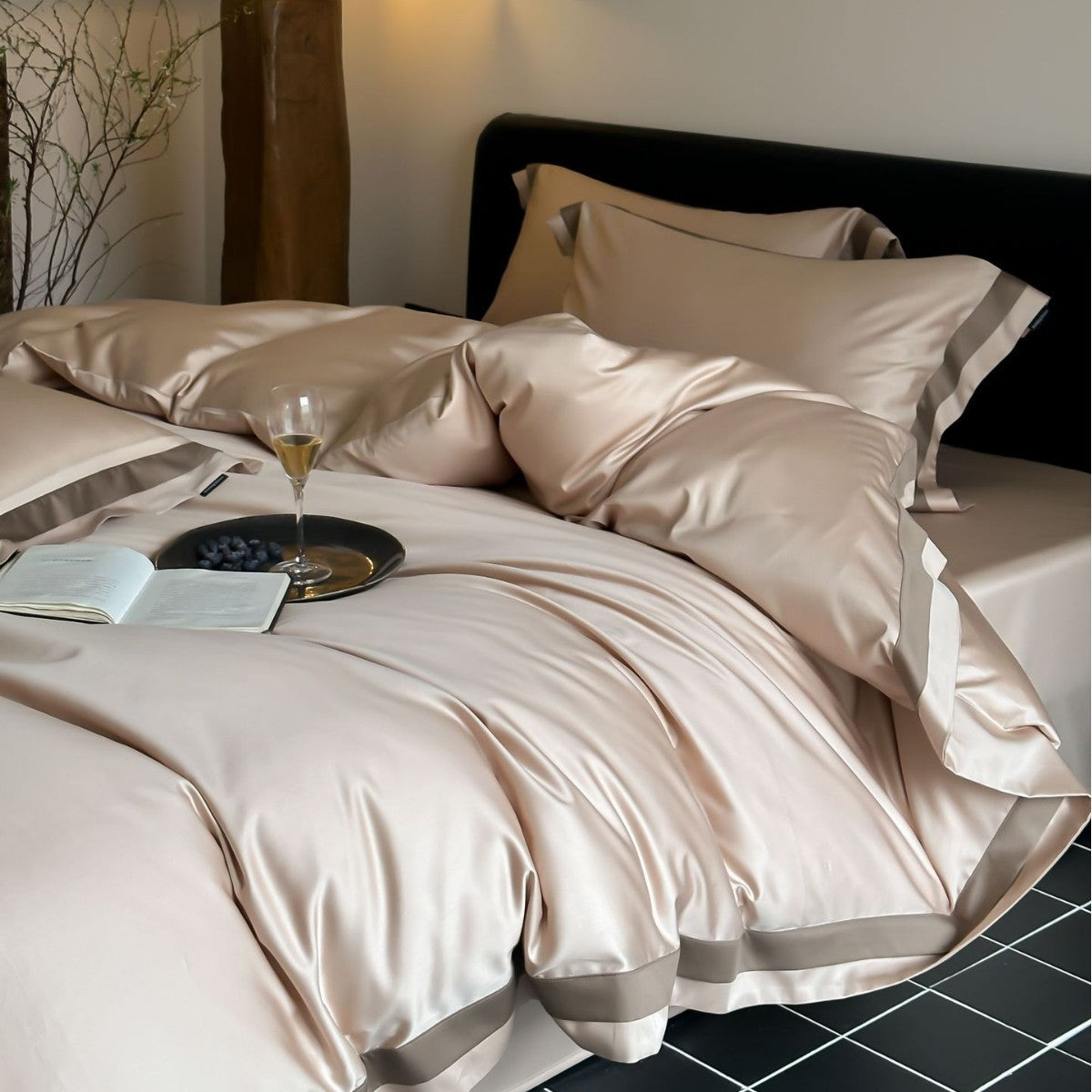 Neatly made bed with beige satin sheets and pillows, accompanied by a glass of wine and book.