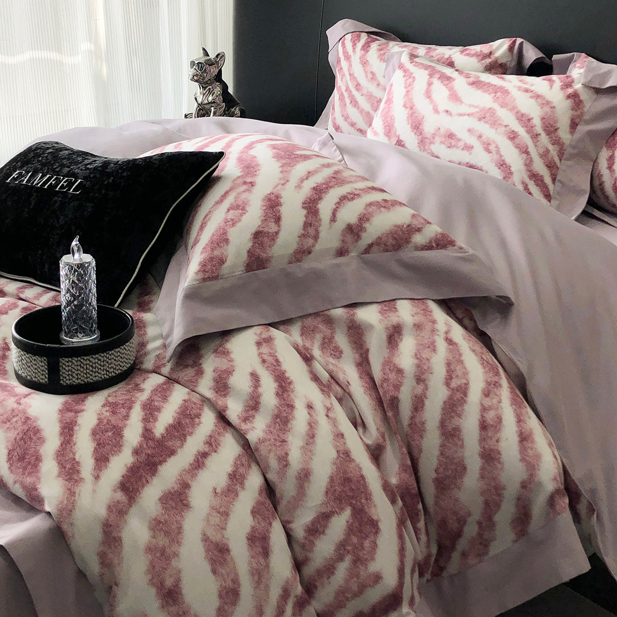 Bed with pink and white patterned bedding, gray pillows, and a small cat on a white curtain background.