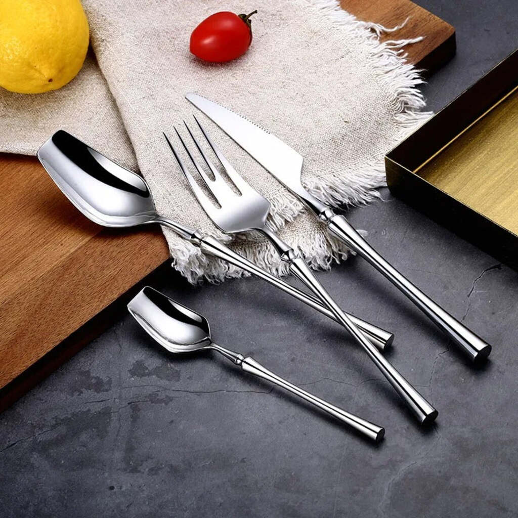 Set of silver cutlery on a dark surface with a wooden cutting board and fruits in the background.