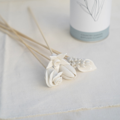 White flower diffuser sticks on a light gray surface with a bottle in the background