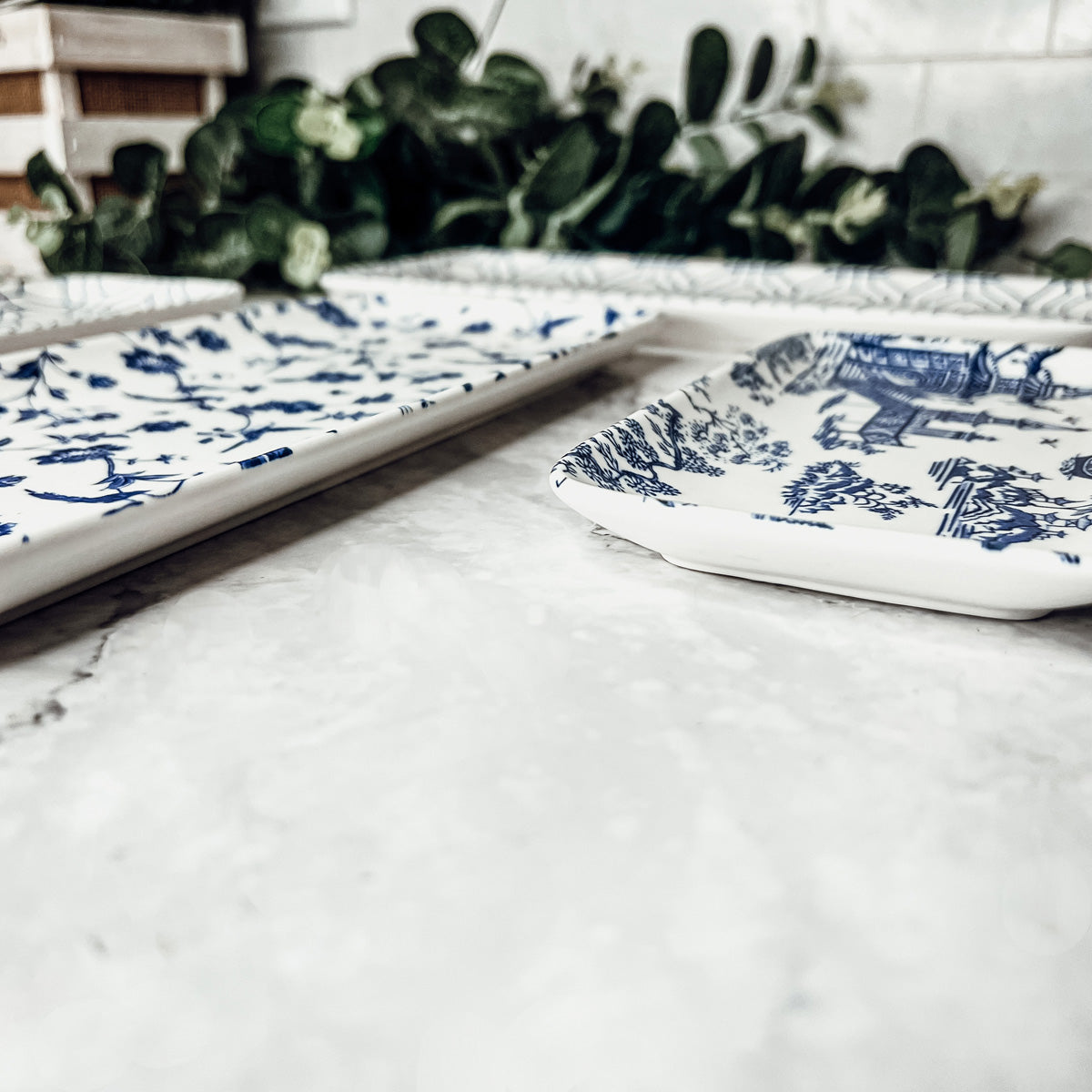 Two white trays with blue floral patterns on a marble surface, with green plants in the background.