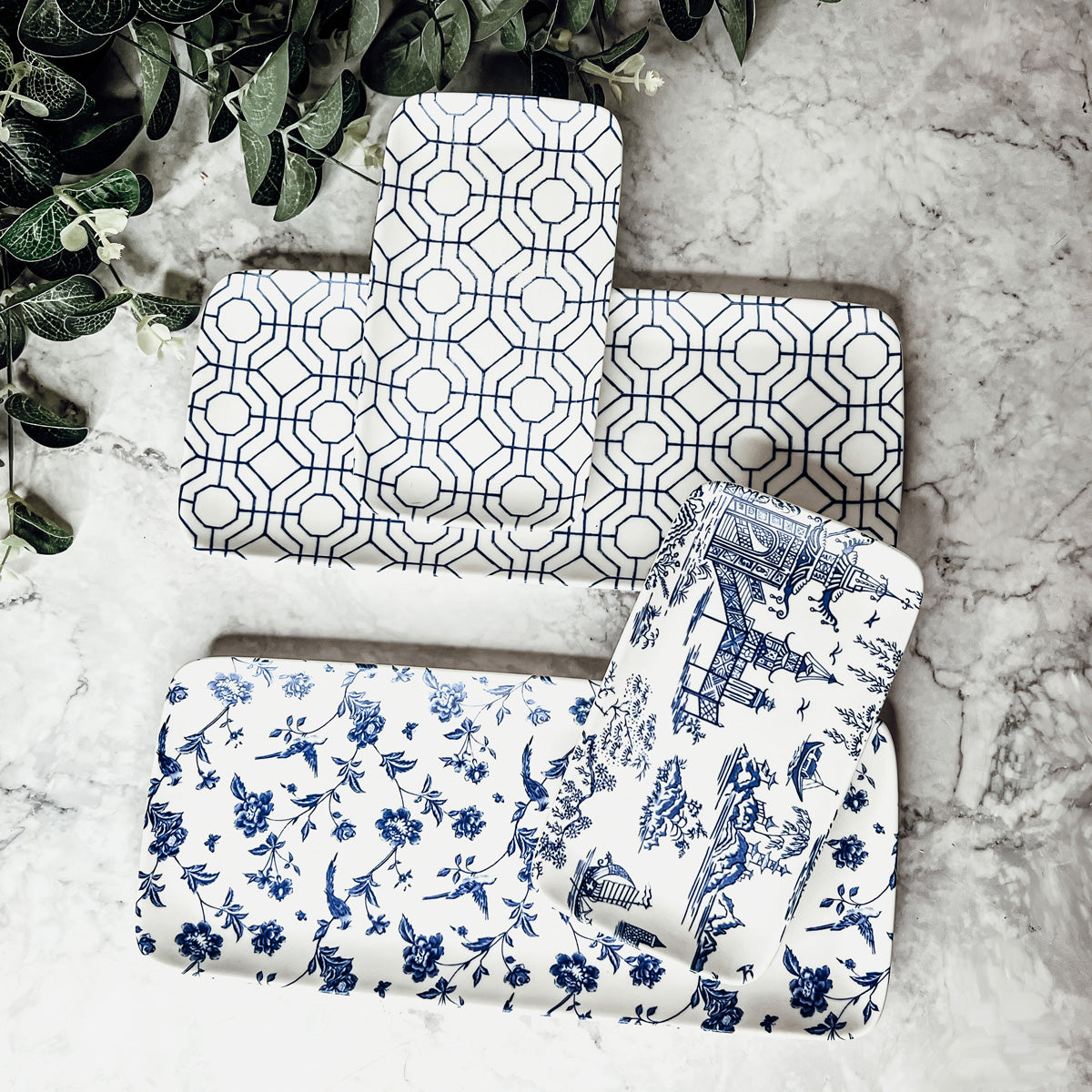 Chinoiserie and geometric serving trays on a marble surface with greenery.