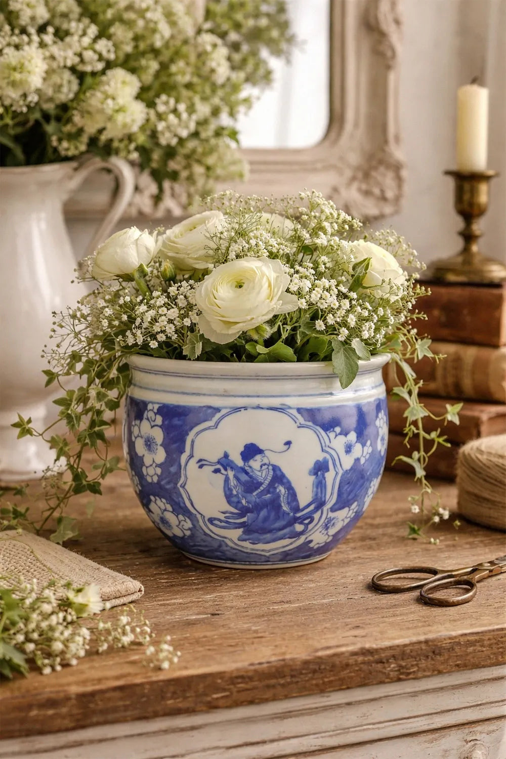 Floral arrangement in a blue and white chinoiserie ceramic pot on a wooden surface with a mirror and candle in the background.