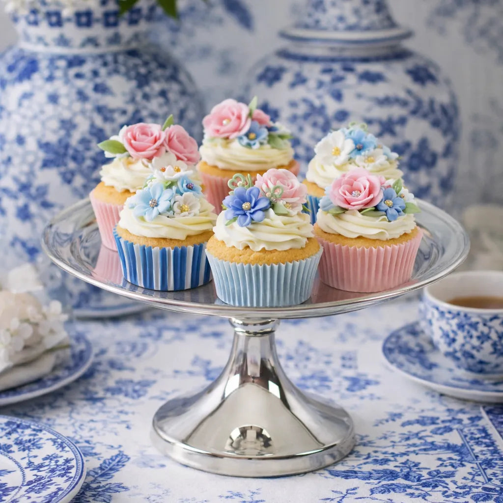Decorative cupcakes with flowers on a silver-plated Tazza stand against a blue and white floral tablecloth.