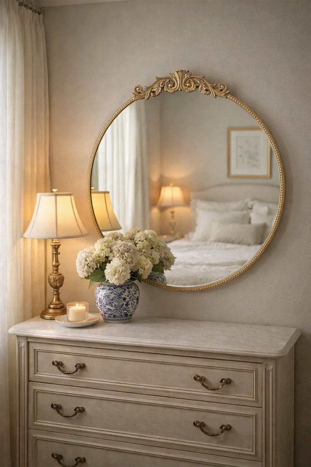 Decorative round mirror on a dresser with floral arrangement and lamp.
