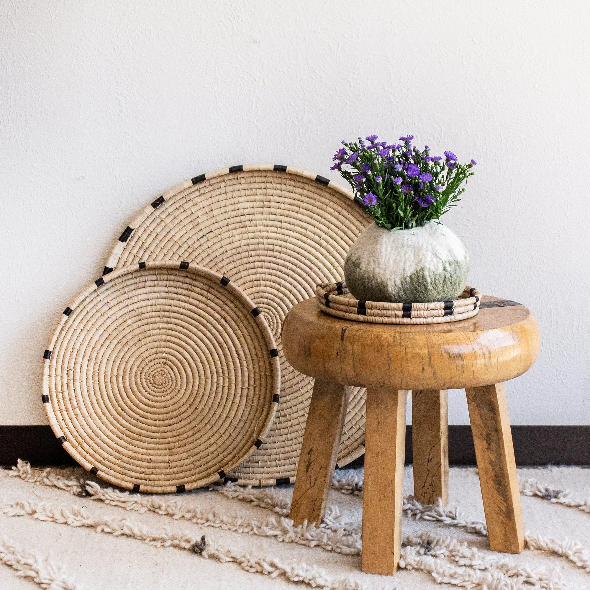 Woven decorative trays and a wooden stool with a vase of flowers on a white wall background