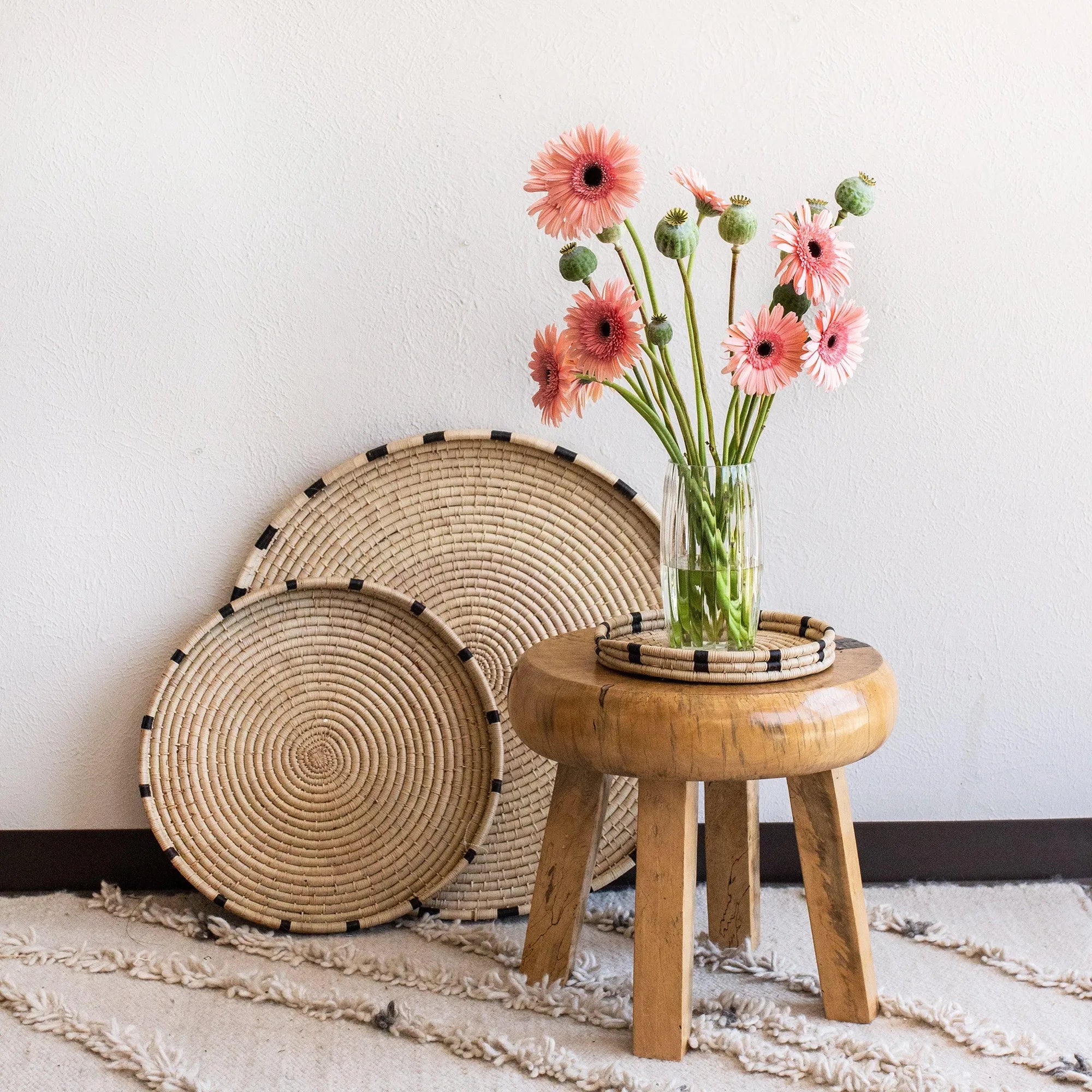 Wooden stool with a vase of pink flowers and decorative woven trays against a white wall.