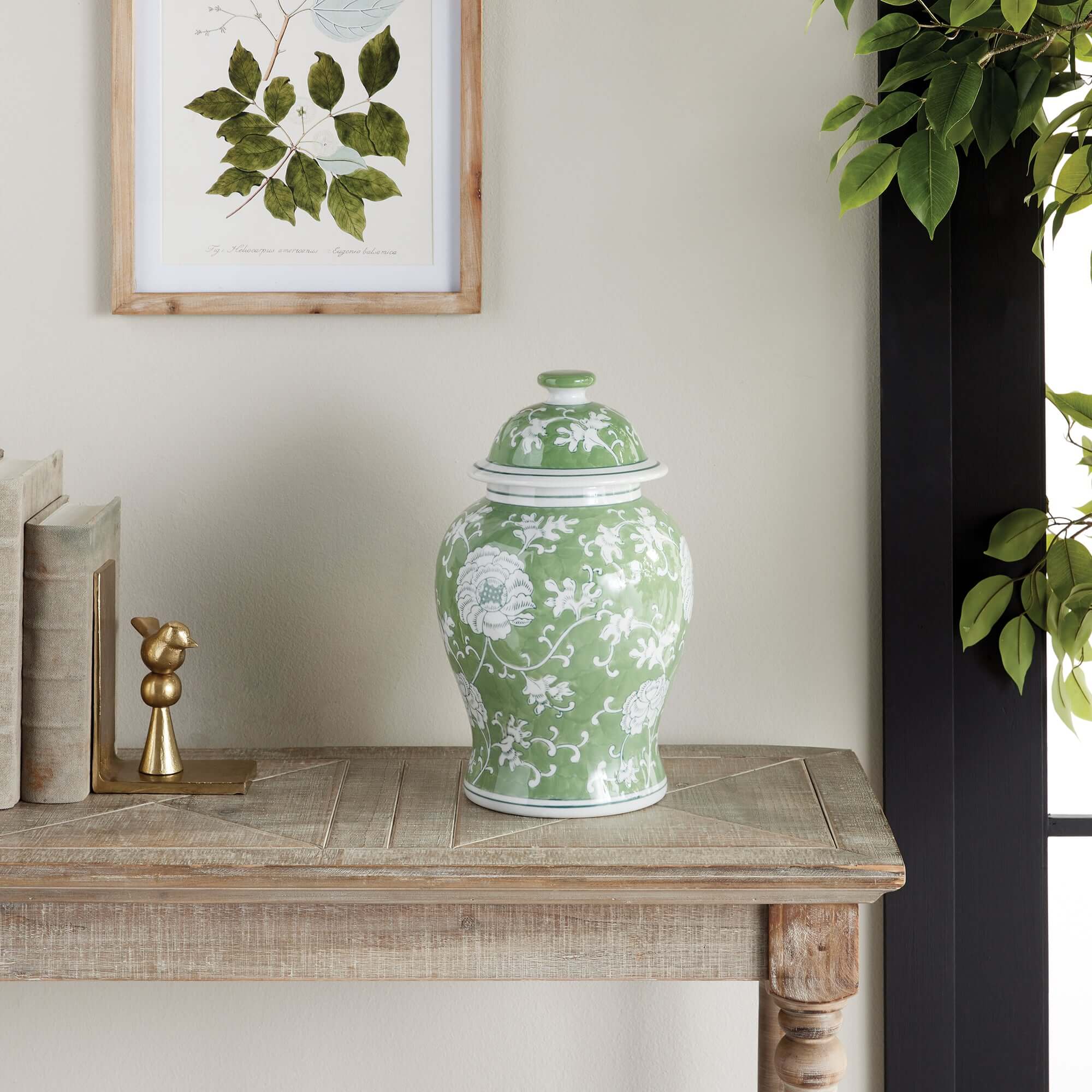 Green ceramic jar with floral patterns on a wooden table, framed artwork in the background