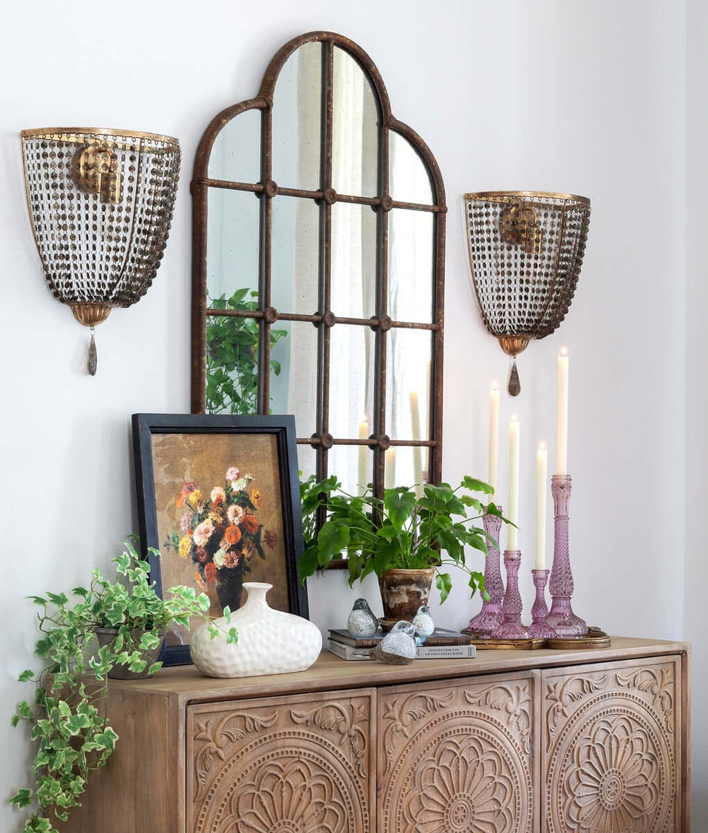 Decorative wall setup with a large mirror, wall sconces, candles, and plants on a wooden console table.