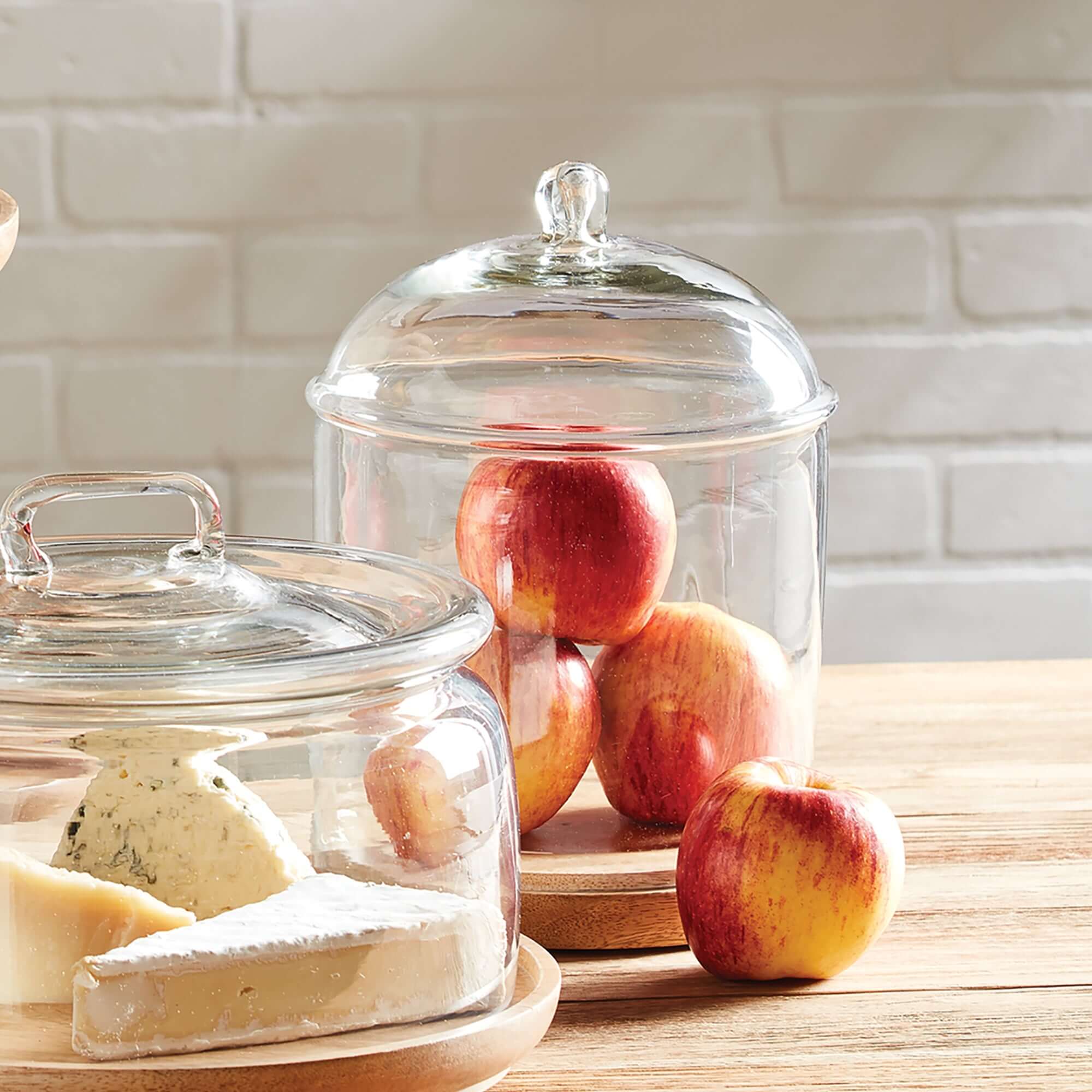 Glass cloches with lids on a wooden surface, containing apples and cheese.