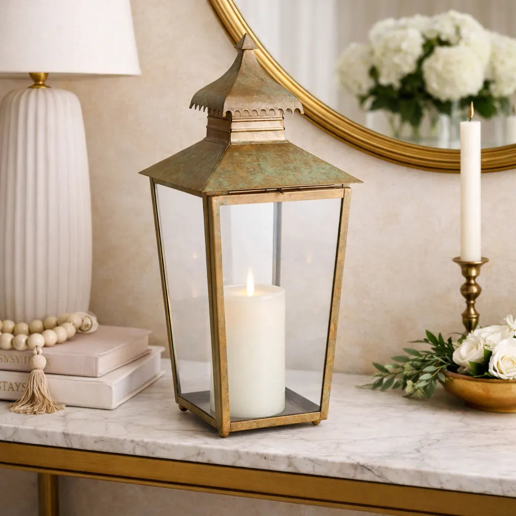 Decorative metal lantern with a candle on a marble surface, with a mirror and flowers in the background.
