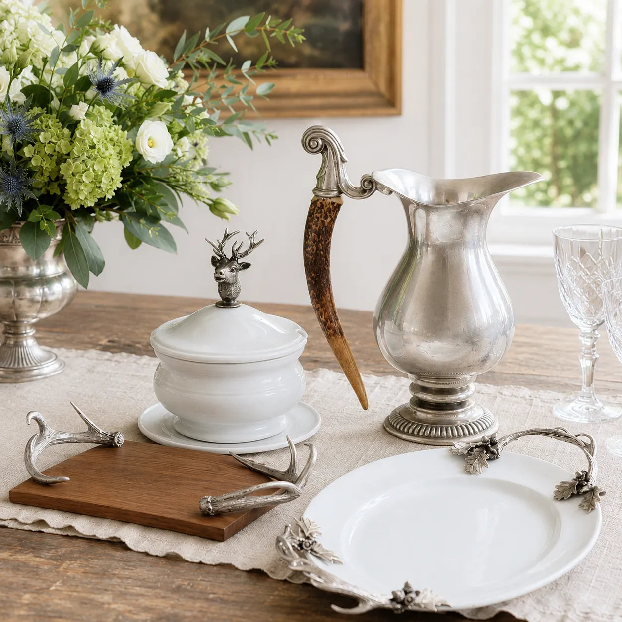 A styled tabletop arrangement featuring a silver pitcher with a curved antler handle, a white lidded ceramic bowl with a deer-shaped knob, a white serving platter with ornate silver handles, a wooden board, and floral decor in the background.