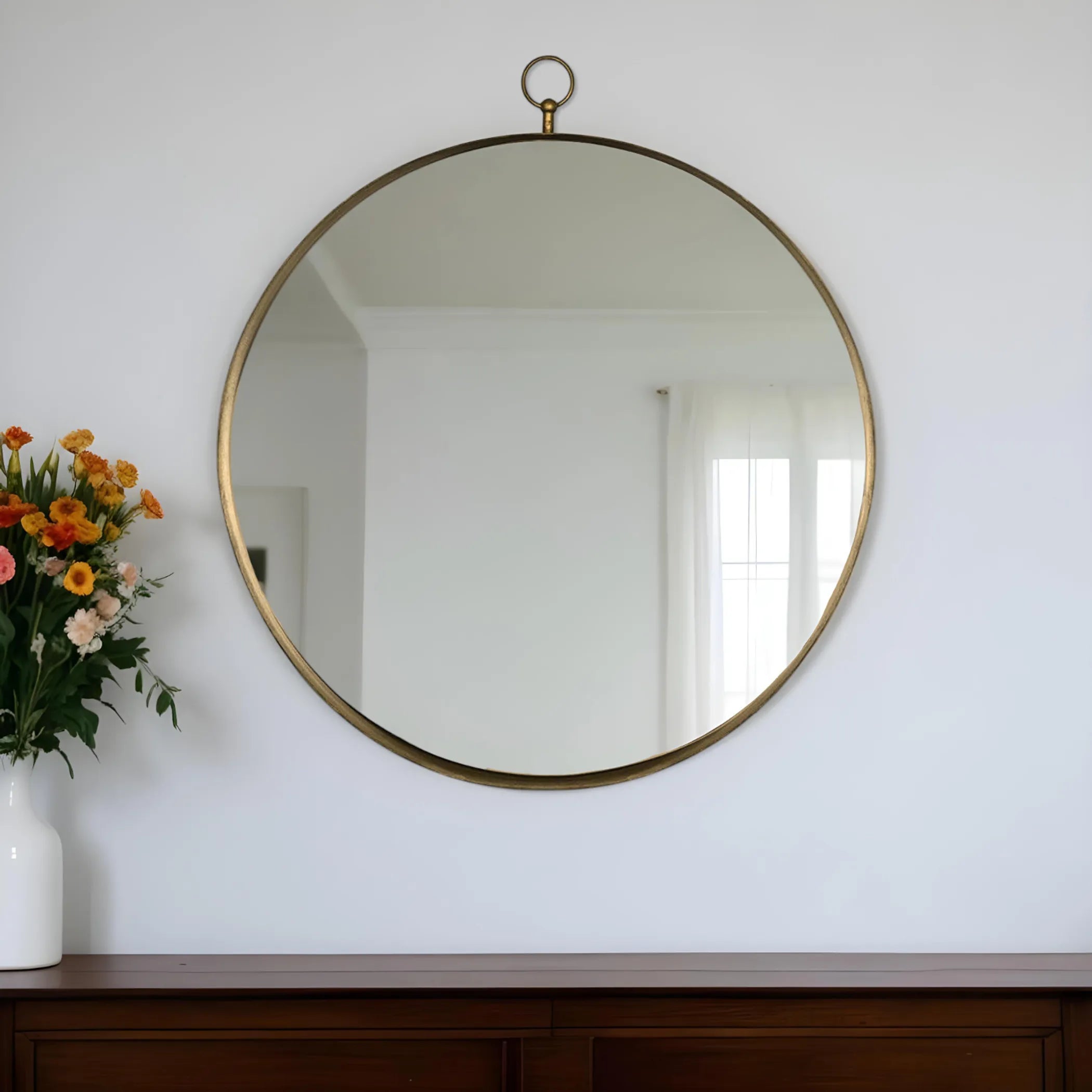 Round gold-framed mirror on a white wall with a vase of flowers to the left.