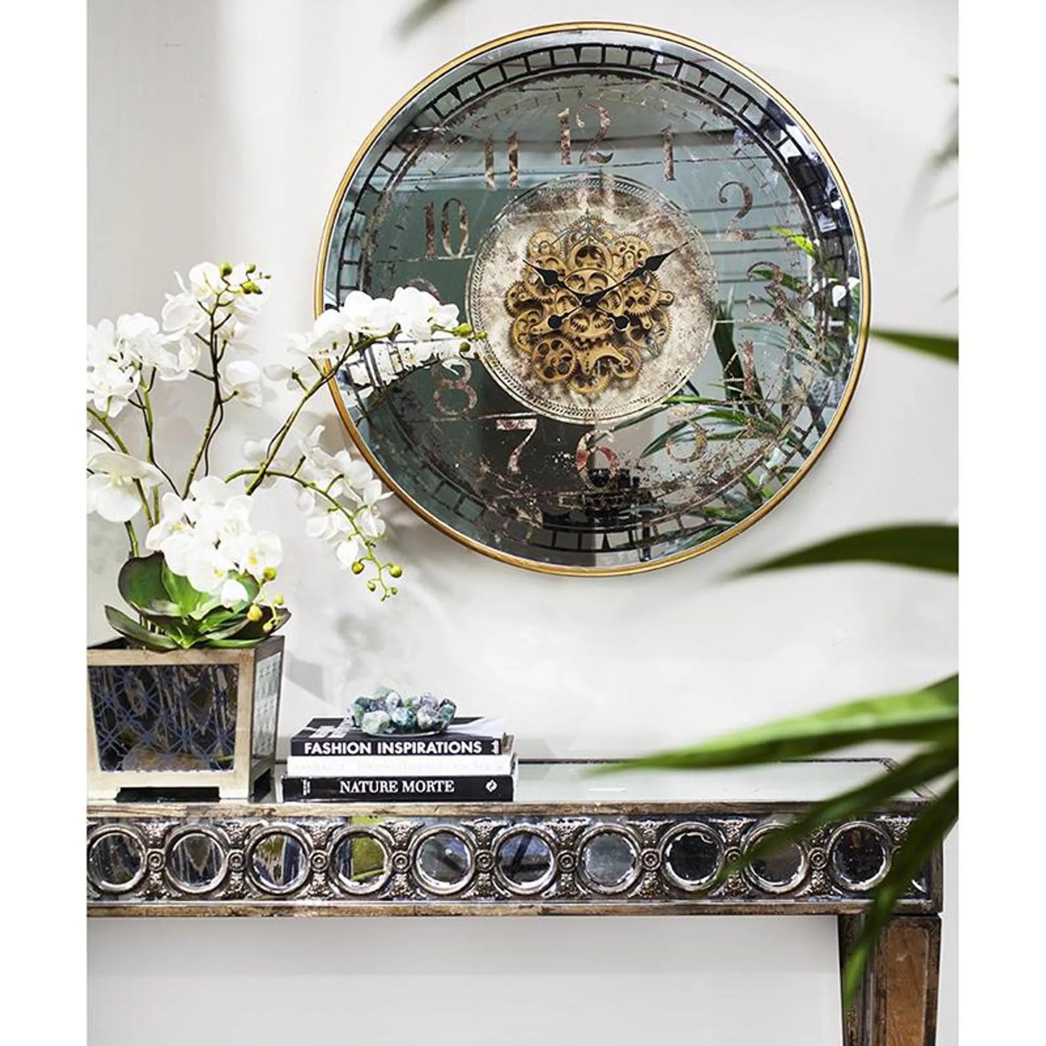Decorative wall clock with intricate design on a white wall, with a plant and books in the foreground.