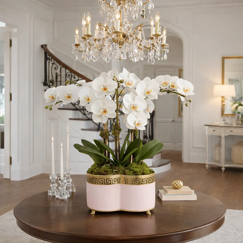 White orchids in a decorative pink planter with greek key design and gold feet on a wooden table with a chandelier in the background.
