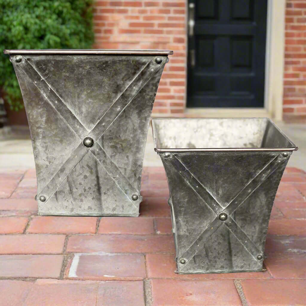 Two metal planters on a brick patio with a brick wall and door in the background.