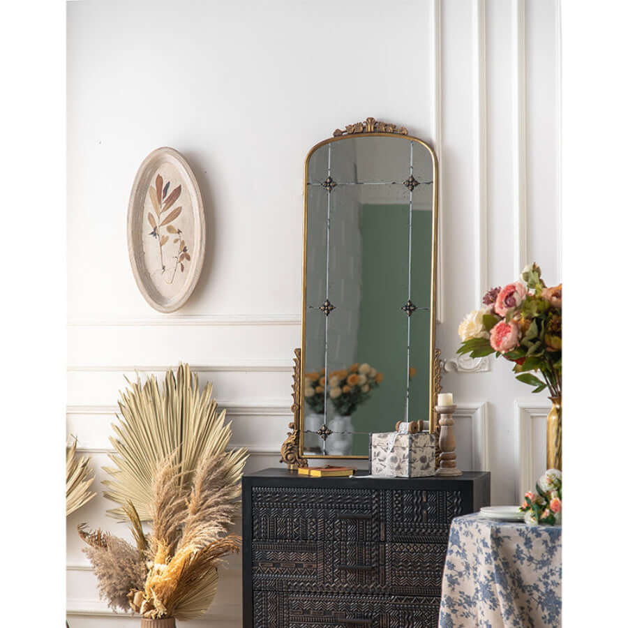 Decorative interior with a tall mirror, framed artwork, and floral arrangements.