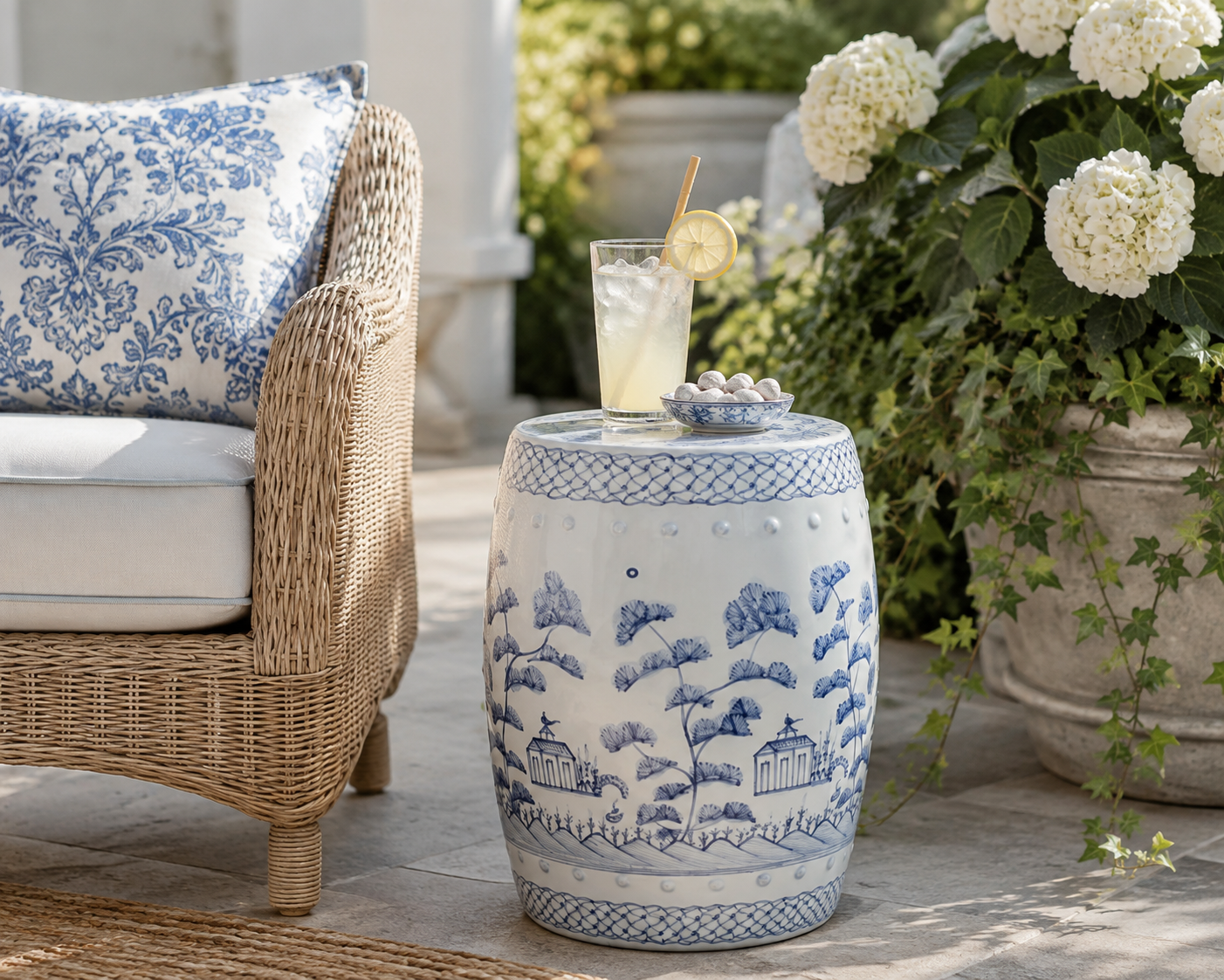 A blue and white porcelain garden barrel side table styled outdoors next to a wicker chair with a blue and white patterned pillow, holding a glass of lemonade and a small bowl of marbles.