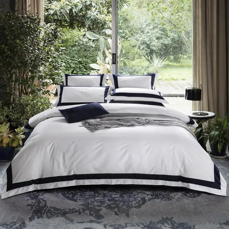 Neatly made bed with white bedding and black accents in a room with large windows and greenery.