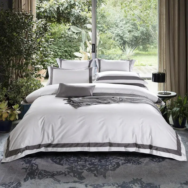 Neatly made bed with white bedding and gray accents in a room with large windows and greenery.