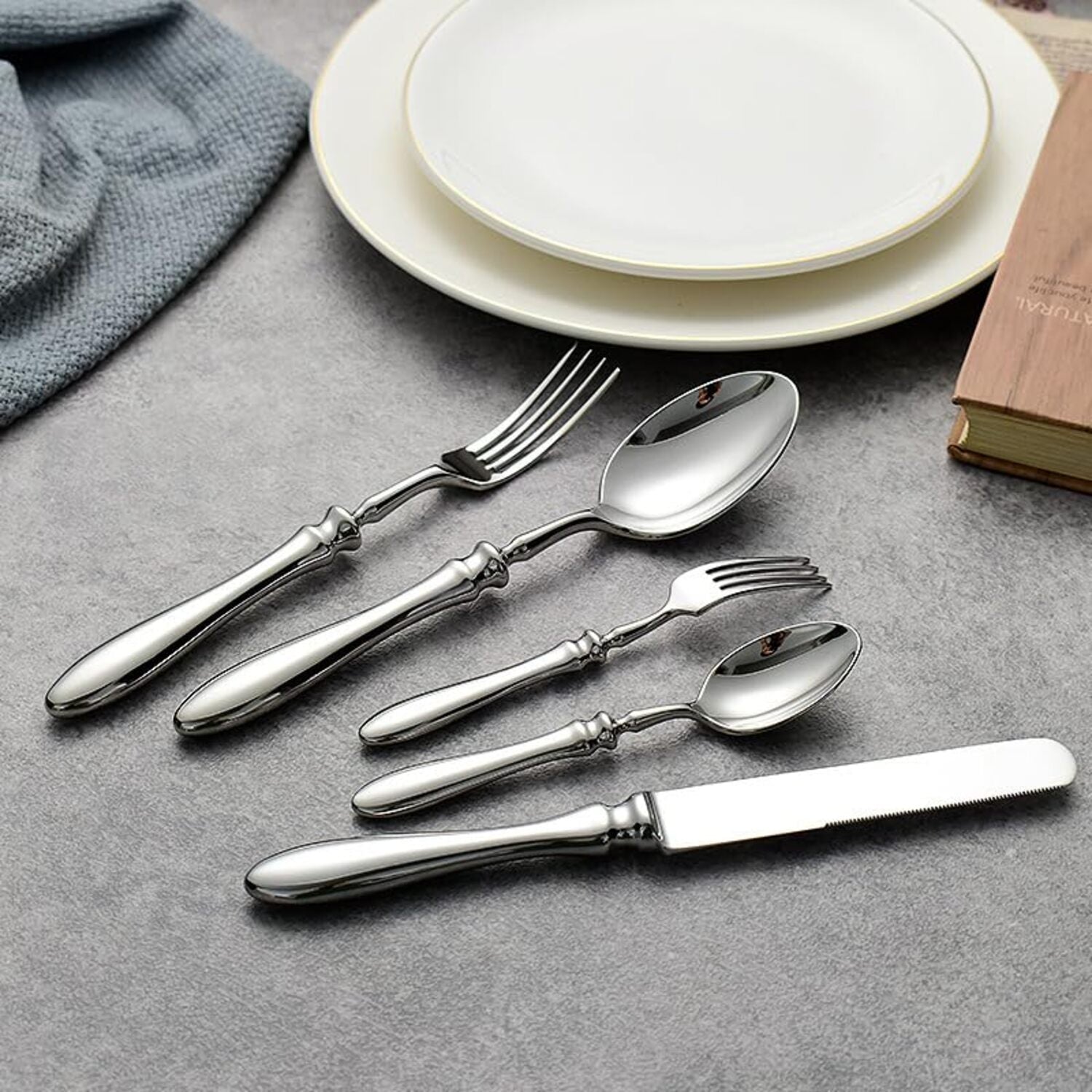 Set of silver cutlery including forks, knives, and spoons on a gray surface with plates in the background.