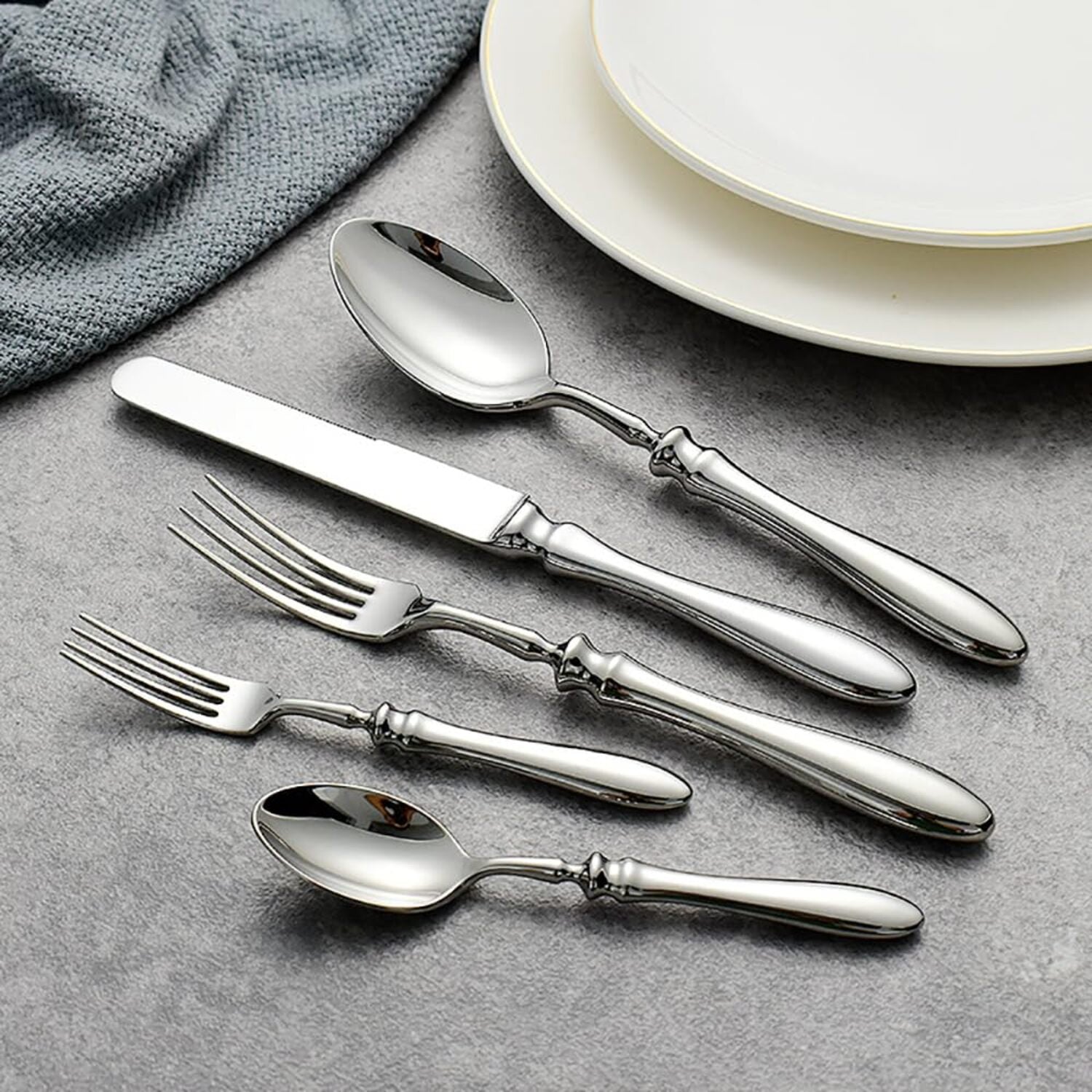 Set of silver cutlery including forks and spoons on a gray surface with plates in the background.