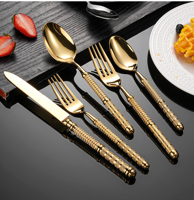 Set of gold cutlery on a reflective surface with fruit and a plate in the background