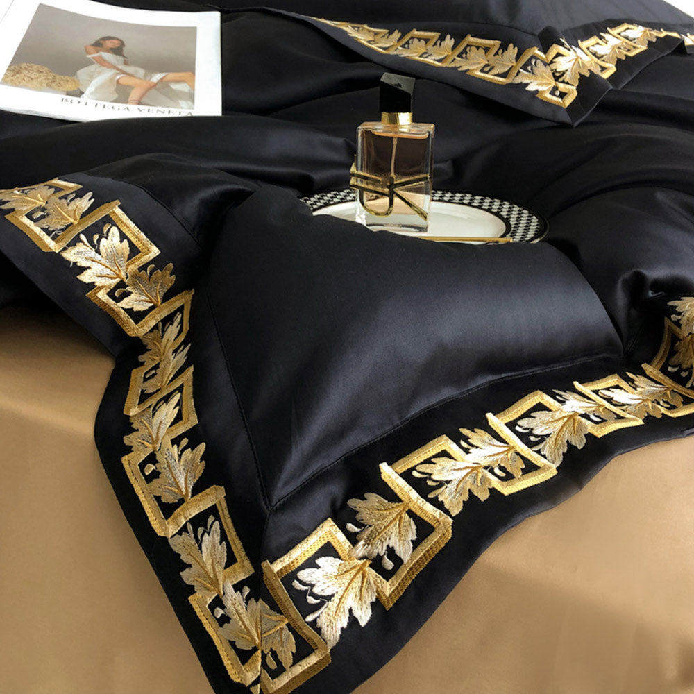 Black satin pillow with gold embroidery on a beige surface, next to a magazine and perfume bottle.