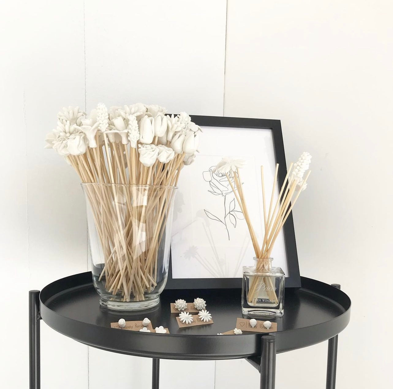 Decorative white flower diffuser sticks, and a framed picture on a black round table.