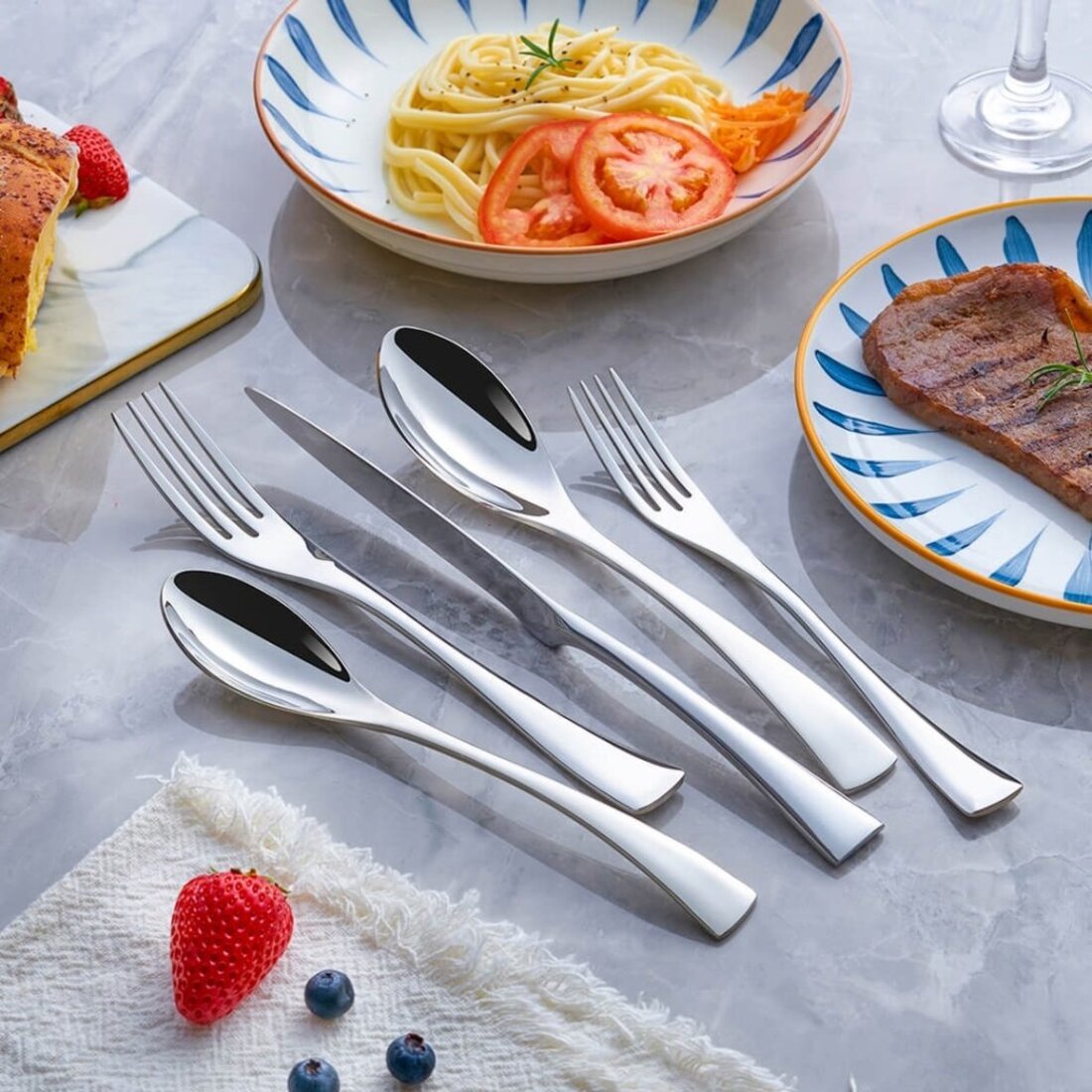 Set of silver cutlery on a table with plates of food including pasta and toast.