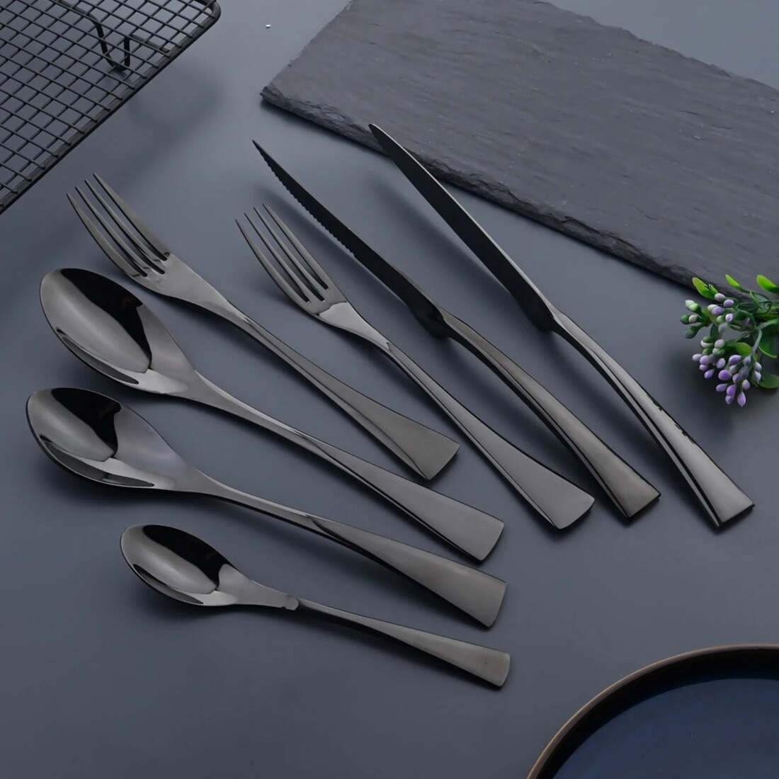 Set of black cutlery including forks, knives, and spoons on a dark surface with a plant.