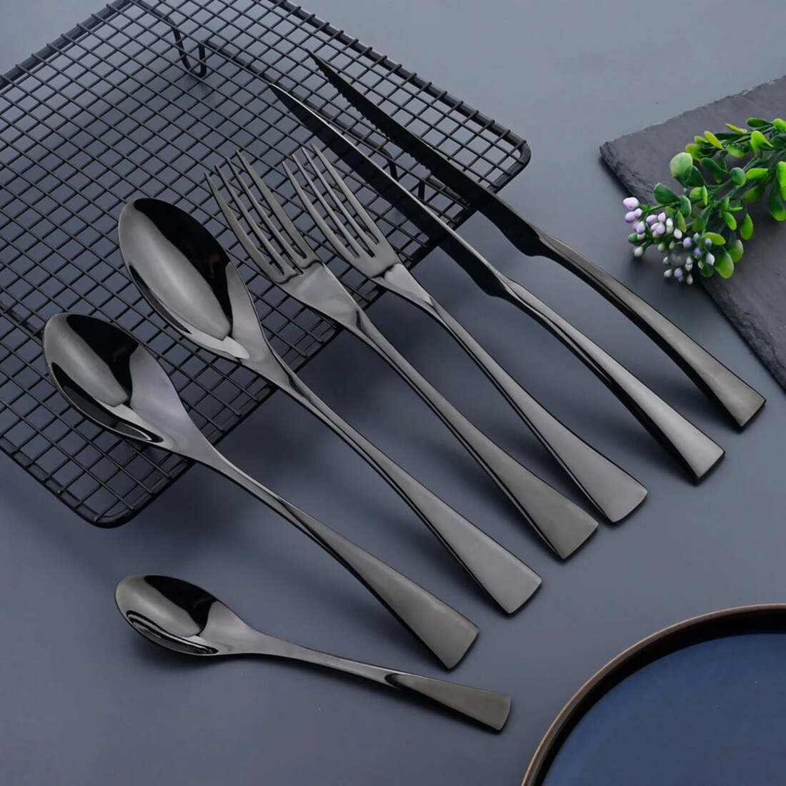 Set of black cutlery including knives, forks, and spoons on a dark surface with a plant in the corner.
