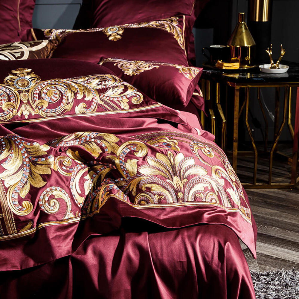 Luxurious burgundy and gold embroidered bedding set on a bed with a side table in the background.